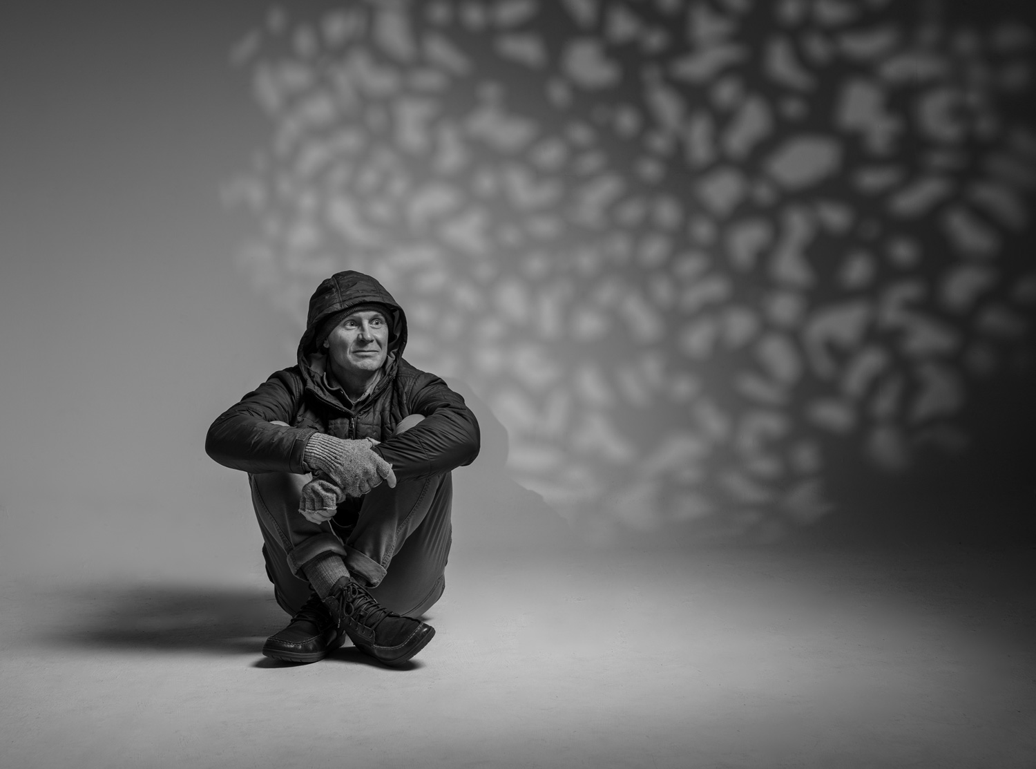 Black-and-white portrait of a man sitting on the floor hugging his knees in a hooded jacket, with dappled leaf-shadow light on the wall behind him