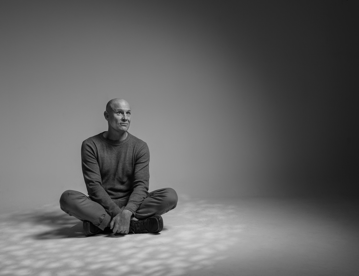 Black-and-white portrait of a man sitting cross-legged and looking upward, with dappled shadow patterns cast around him