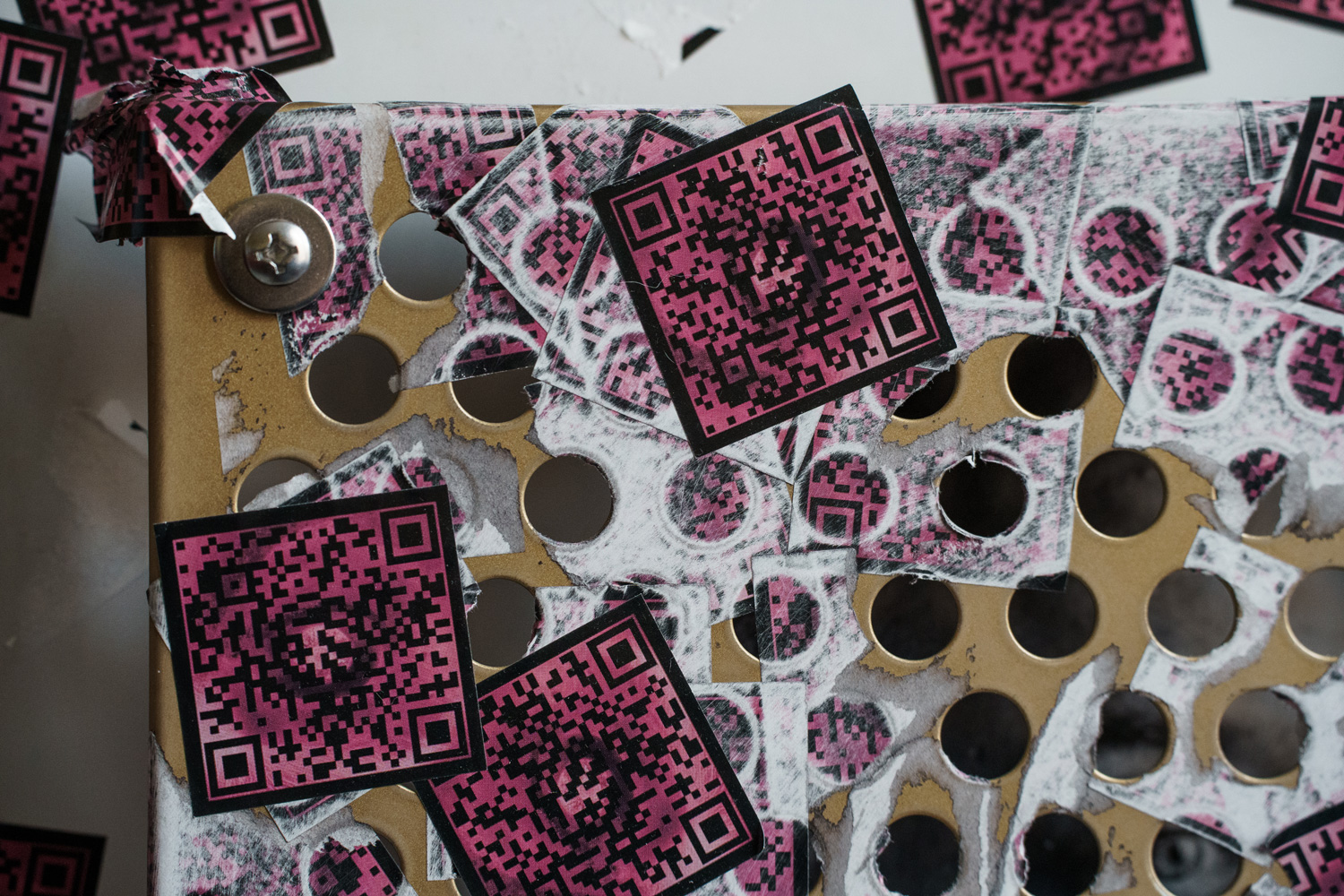Detail of a torn pink QR code sticker peeling at the corner of the perforated gold panel, revealing a screw beneath