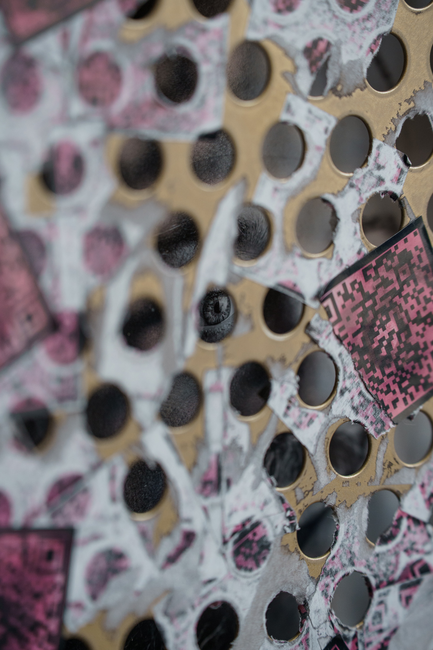 Macro detail of the perforated gold panel with a human eye visible through one of the holes, framed by out-of-focus torn QR code stickers