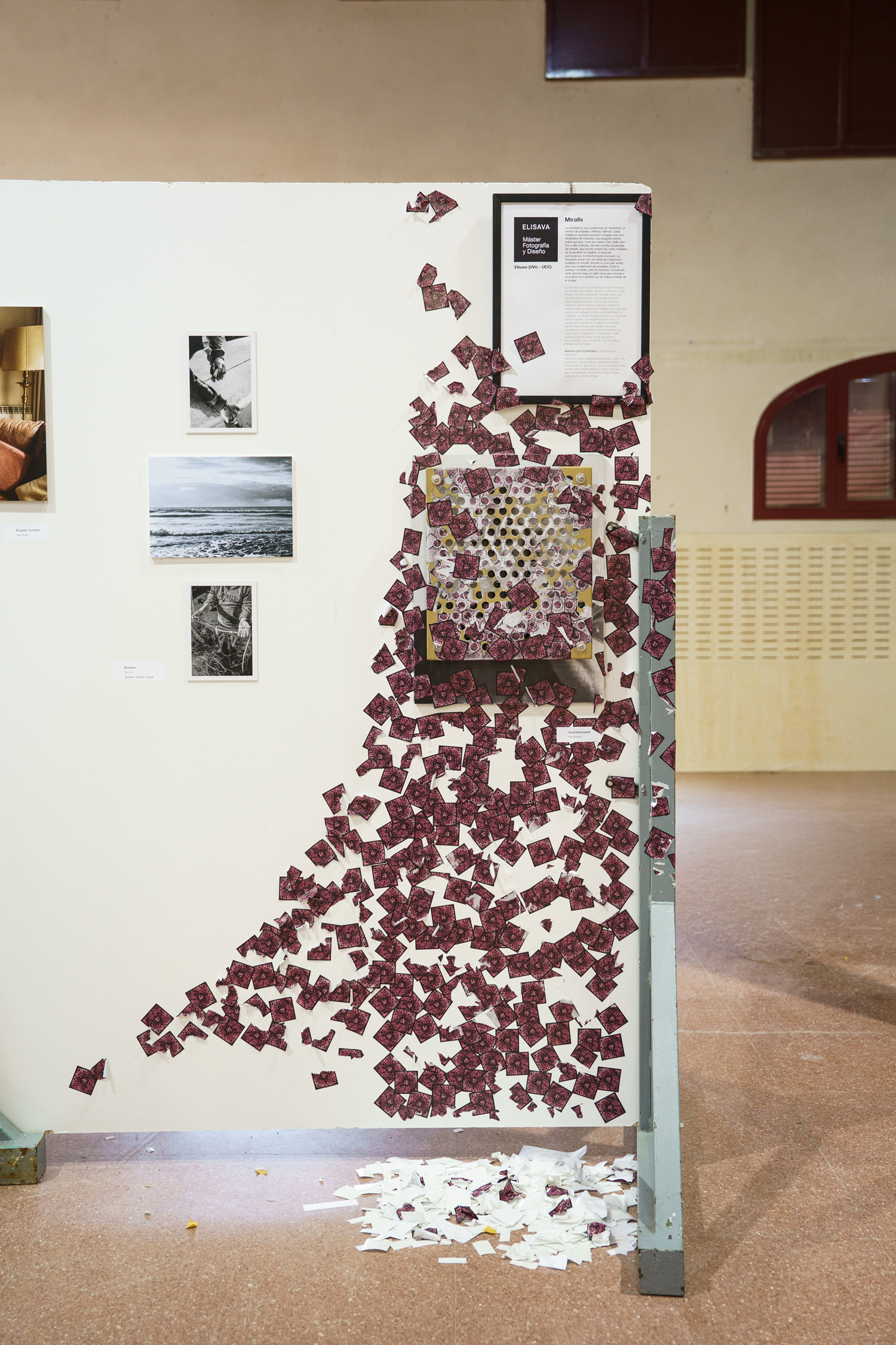 Gallery wall showing black-and-white portrait photographs beside color photo diptychs, with the Overstimulated mixed-media piece — a perforated gold panel plastered with torn pink QR code stickers cascading down the wall and onto the floor